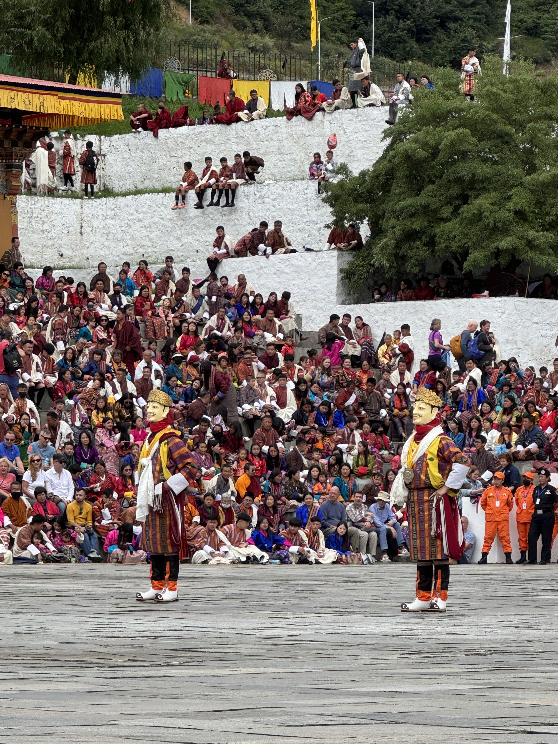 Two masked cham dancers in gold brocade performing before a seated crowd in national dress at a Bhutanese tshechu